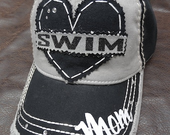 May include: Gray and black baseball cap with a black heart patch that says "SWIM" in white letters. The brim of the cap has the word "Mom" written in white letters.
