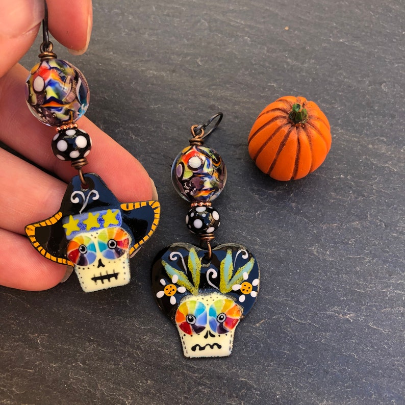 Puede incluir: Un par de pendientes coloridos hechos a mano con dijes de calavera de az&uacute;car con detalles en blanco y negro. Los pendientes est&aacute;n hechos con cuentas de vidrio y alambre de cobre.