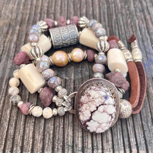 May include: A collection of beaded bracelets in shades of cream, purple, and silver. The bracelets feature various beads, including pearls, cylindrical bone-like beads, and a large oval pendant with a marbled design. A brown suede tassel is attached.