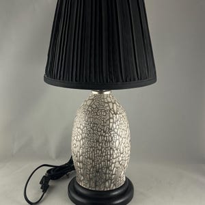 May include: A decorative table lamp with a black pleated lampshade. The lamp base has a white and gray crackle glaze pattern, and sits on a black wooden base. The lamp includes a black power cord.