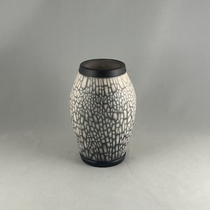 May include: A ceramic vase with a cylindrical shape. The vase has a white base with a black, crackled pattern. The top and bottom rims are solid black. The interior of the vase is dark.