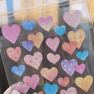 May include: A sheet of colorful heart-shaped masking stickers. The stickers have different patterns, including polka dots, stripes, and plaid. The sheet is packaged in a clear plastic bag with the text "MASKING STICKER" and "ADESIVO" printed on it.