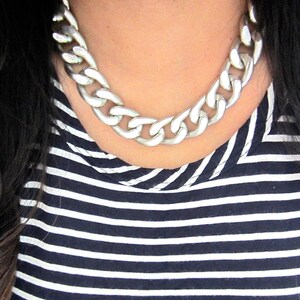 May include: A silver chunky chain necklace with large links. The necklace is worn by a person with dark hair and a striped shirt.