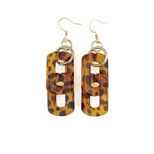 May include: A pair of gold tone dangle earrings with a leopard print design. The earrings are made of two linked rectangular shapes.