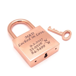 以下が含まれることがあります： ローズゴールドの南京錠と鍵のセット。南京錠には「M&D Locked in Love」、日付「9.2.2017」、座標「33.9526° N 84.5499° W」が刻印されています。滑らかな金属仕上げで、標準的なU字型のシャックルが付いています。