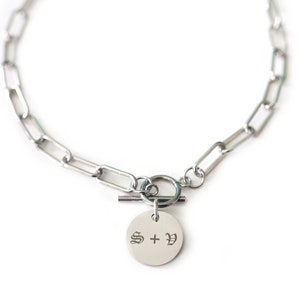 May include: Silver chain necklace with a toggle clasp and a round pendant engraved with "S + V" in a gothic font.