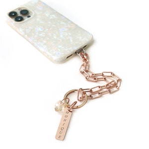 May include: A white phone case with a rose gold chain and a charm with the name "MONIQUE" engraved on it.