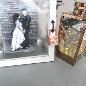 May include: A rose gold padlock with the inscription "M&D Locked in Love 9.2.2017 13.05.10 84.5499" hanging from a white frame with a black and white photo of a couple inside. The padlock is hanging in front of a rose gold lantern with string lights inside.