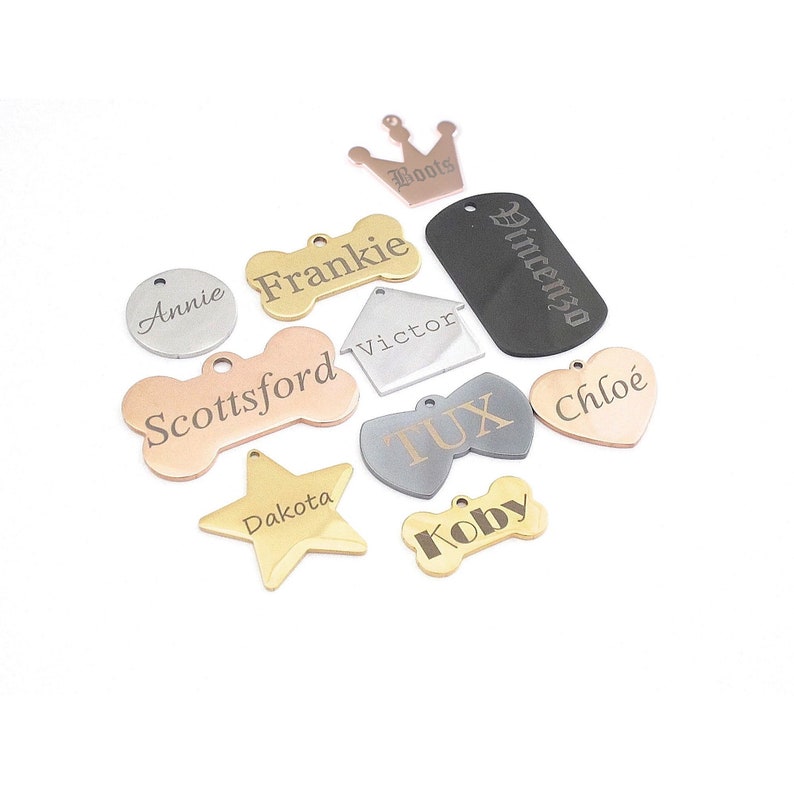 May include: A collection of personalized pet ID tags in various shapes and colors. The tags include a gold star, a silver circle, a heart-shaped tag, and bone-shaped tags. Each tag is engraved with a name, such as "Annie", "Frankie", and "Dakota".