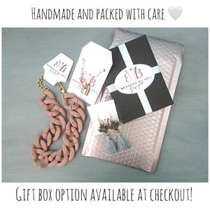 May include: An assortment of jewelry, including a chunky pink necklace, a gold chain necklace with a lock pendant, and earrings with gray tassels. The items are packaged in a black box and pink bubble mailer. Text reads "HANDMADE AND PACKED WITH CARE".