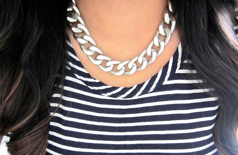 May include: A silver chunky chain necklace with large links. The necklace is worn by a person with dark hair and a striped shirt.