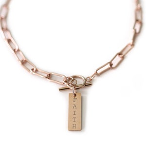 Op de afbeelding: Een gouden ketting met een toggle-sluiting en een rechthoekige hanger met de tekst "FAITH" in verticale letters.