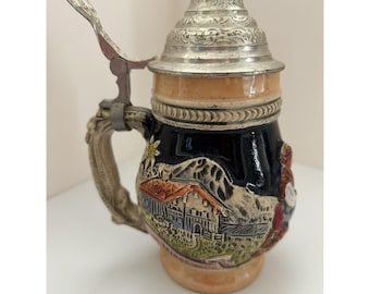 May include: A ceramic beer stein featuring a dark blue body with a raised, colorful relief depicting a mountain landscape, a house, and a figure. It includes a metallic, hinged lid with intricate engravings and a curved, textured handle.