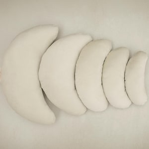 May include: A set of five off-white, crescent-shaped pillows of varying sizes, arranged in a row. The pillows are made of a soft, plush material. A small tag is visible on the largest pillow. The pillows are designed for comfort and support.