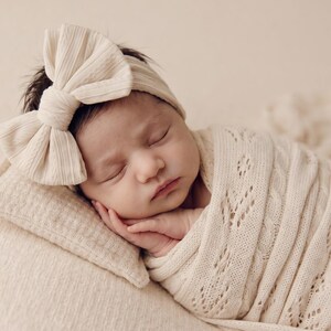 May include: A sleeping infant wrapped in a cream-colored knit swaddle blanket with a delicate leaf pattern. The infant wears a matching cream-colored ribbed headband featuring a large, prominent bow detail positioned on the side of the head.