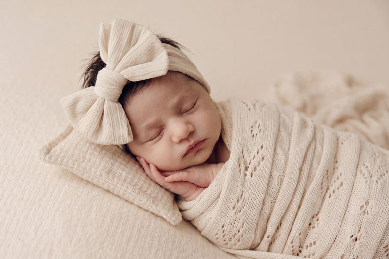 May include: A sleeping infant wrapped in a cream-colored knit swaddle blanket with a delicate leaf pattern. The infant wears a matching cream-colored ribbed headband featuring a large, prominent bow detail positioned on the side of the head.