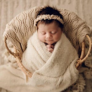May include: A newborn infant with dark hair is wrapped in a soft, cream-coloured fuzzy blanket and placed inside a round woven wicker basket. The infant wears a delicate, textured cream-coloured headband and has eyes closed in a peaceful expression.