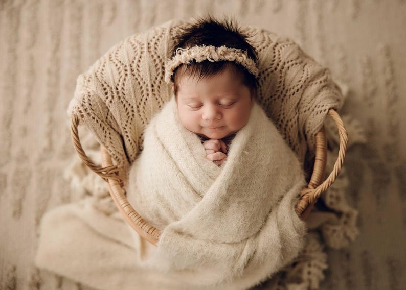 May include: A newborn infant with dark hair is wrapped in a soft, cream-coloured fuzzy blanket and placed inside a round woven wicker basket. The infant wears a delicate, textured cream-coloured headband and has eyes closed in a peaceful expression.
