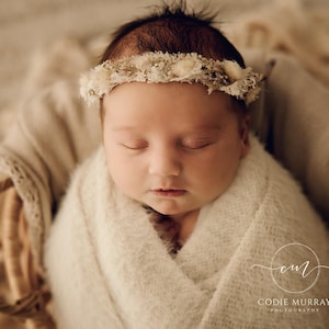 May include: A newborn infant wrapped in a soft, cream-coloured textured blanket, resting inside a woven wicker basket. The infant wears a delicate floral headband featuring small, dried white and beige blossoms and foliage across the forehead.