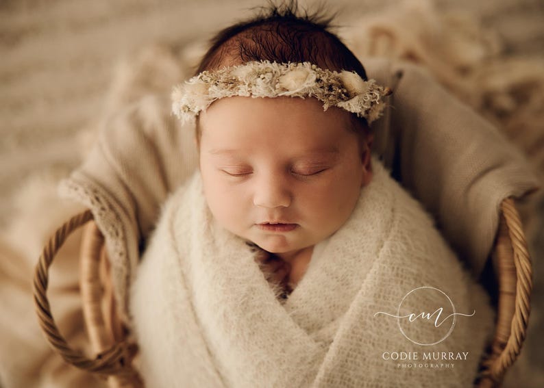 May include: A newborn infant wrapped in a soft, cream-coloured textured blanket, resting inside a woven wicker basket. The infant wears a delicate floral headband featuring small, dried white and beige blossoms and foliage across the forehead.