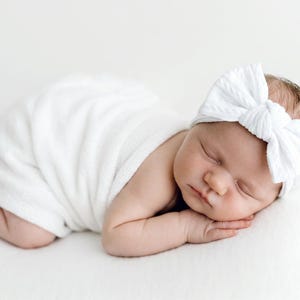 May include: A newborn infant is wrapped in a soft white swaddle blanket and wears a white ribbed headband featuring a large, centered bow. The infant is lying in a prone position with eyes closed, resting on a plain, solid white surface.