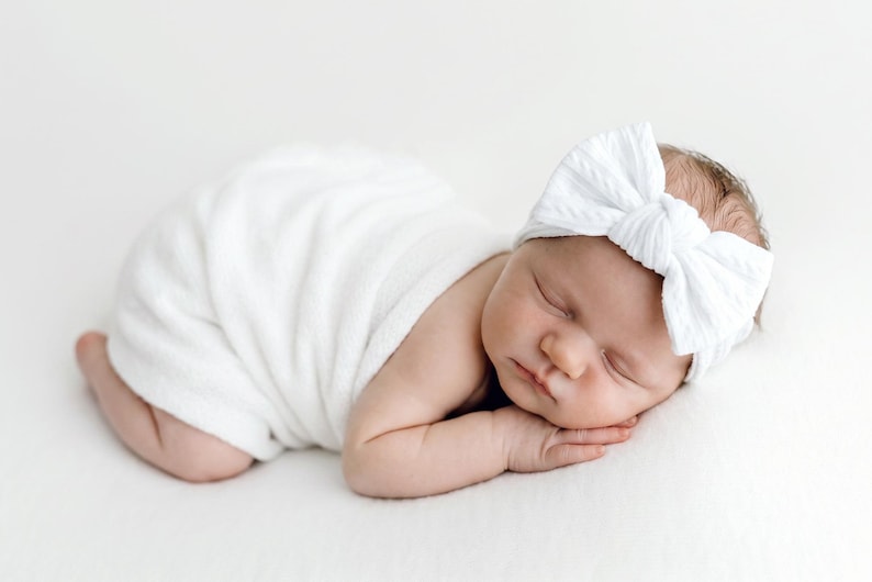 May include: A newborn infant is wrapped in a soft white swaddle blanket and wears a white ribbed headband featuring a large, centered bow. The infant is lying in a prone position with eyes closed, resting on a plain, solid white surface.
