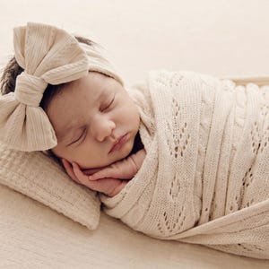 May include: A sleeping infant wrapped in a cream-colored knit swaddle blanket with a delicate leaf-patterned texture. The infant wears a matching cream-colored headband featuring a large, prominent bow detail on the side of the head.
