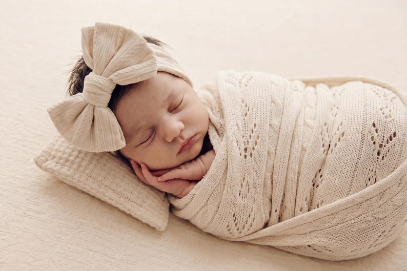 May include: A sleeping infant wrapped in a cream-colored knit swaddle blanket with a delicate leaf-patterned texture. The infant wears a matching cream-colored headband featuring a large, prominent bow detail on the side of the head.