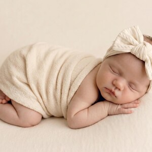 May include: A sleeping infant wrapped in a cream-colored swaddle blanket, wearing a matching cream-colored headband with a large, textured bow detail. The fabric of both the swaddle and the headband features a soft, ribbed, and knit-like texture.