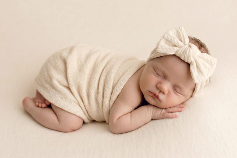 May include: A sleeping infant wrapped in a cream-colored swaddle blanket, wearing a matching cream-colored headband with a large, textured bow detail. The fabric of both the swaddle and the headband features a soft, ribbed, and knit-like texture.