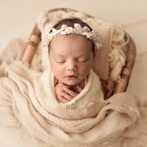 May include: A swaddled newborn baby wearing a floral headband, nestled in a woven basket. The baby is wrapped in a cream-coloured blanket. The image is softly lit, creating a calm and peaceful scene, perfect for newborn photography.