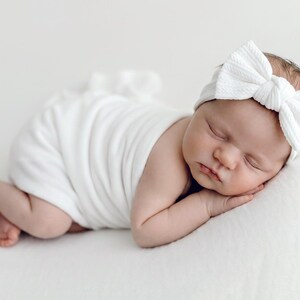 May include: A white ribbed fabric headband featuring a large, centered bow detail, worn by a sleeping infant wrapped in a matching white swaddle blanket. The headband has a textured, knit-like appearance and sits securely across the forehead.