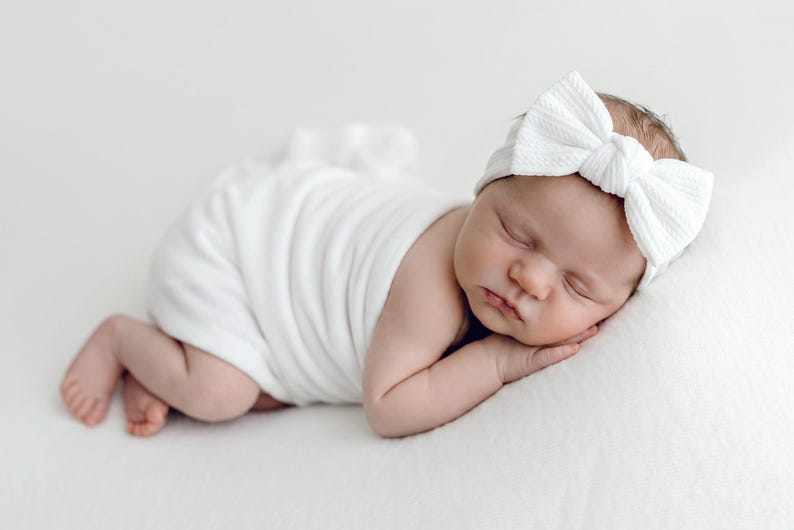 May include: A white ribbed fabric headband featuring a large, centered bow detail, worn by a sleeping infant wrapped in a matching white swaddle blanket. The headband has a textured, knit-like appearance and sits securely across the forehead.