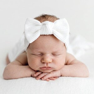 May include: A white ribbed fabric headband featuring a large, centered bow detail. The headband is worn by a sleeping infant who is lying on their stomach with their hands tucked under their chin, resting on a soft white surface.