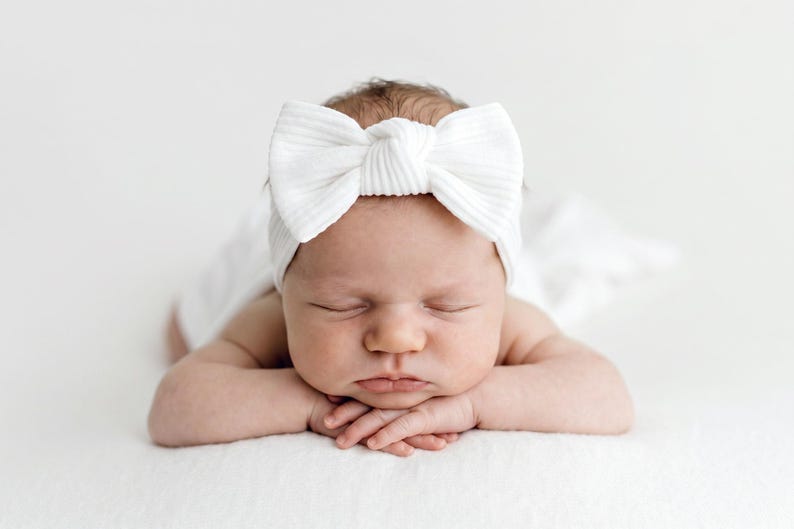 May include: A white ribbed fabric headband featuring a large, centered bow detail. The headband is worn by a sleeping infant who is lying on their stomach with their hands tucked under their chin, resting on a soft white surface.