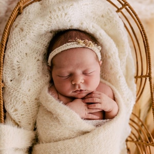 May include: A sleeping newborn wrapped in a cream-coloured textured swaddle blanket, resting inside a curved rattan basket. The infant wears a delicate, thin headband adorned with small, light-coloured floral accents against a soft, fuzzy fabric.