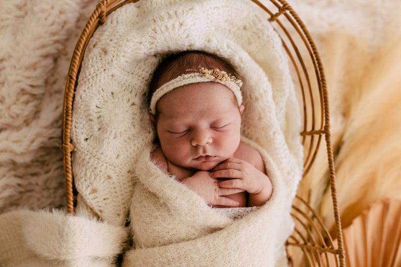 May include: A sleeping newborn wrapped in a cream-coloured textured swaddle blanket, resting inside a curved rattan basket. The infant wears a delicate, thin headband adorned with small, light-coloured floral accents against a soft, fuzzy fabric.