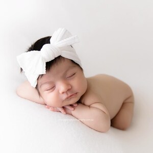 May include: A newborn infant with dark hair is sleeping peacefully while wearing a white fabric headband featuring a large, knotted bow detail on the front. The baby is positioned in a prone pose with arms tucked underneath the chin.