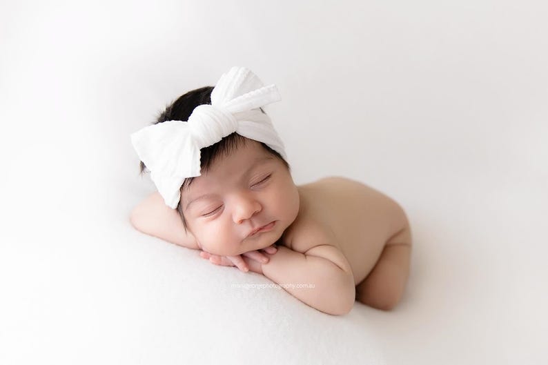 May include: A newborn infant with dark hair is sleeping peacefully while wearing a white fabric headband featuring a large, knotted bow detail on the front. The baby is positioned in a prone pose with arms tucked underneath the chin.