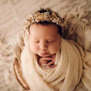 May include: A newborn infant wrapped in a cream-coloured textured swaddle blanket, resting inside a woven basket. The infant wears a decorative headband adorned with small dried flowers, beige grasses, and tiny white beads in a circular arrangement.