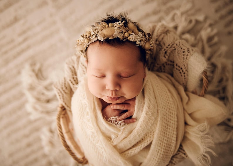May include: A newborn infant wrapped in a cream-coloured textured swaddle blanket, resting inside a woven basket. The infant wears a decorative headband adorned with small dried flowers, beige grasses, and tiny white beads in a circular arrangement.