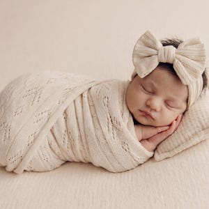 May include: A sleeping infant wrapped in a cream-colored knit swaddle blanket with a delicate openwork pattern. The infant wears a matching cream-colored headband featuring a large, textured fabric bow detail and rests on a small, rectangular pillow.