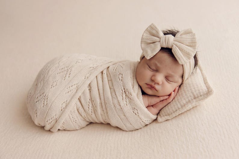May include: A sleeping infant wrapped in a cream-colored knit swaddle blanket with a delicate openwork pattern. The infant wears a matching cream-colored headband featuring a large, textured fabric bow detail and rests on a small, rectangular pillow.
