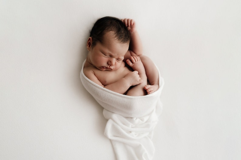 Newborn baby wrapped tightly in white newborn stretch wrap, resting on white blanket. Newborn photography session.