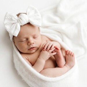 May include: A newborn infant wrapped in a soft white knitted blanket, wearing a large white fabric bow headband. The baby is sleeping with hands tucked near the face, showcasing a textured, ribbed fabric on both the headband and the swaddle wrap.