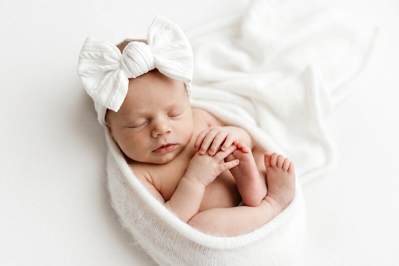May include: A newborn infant wrapped in a soft white knitted blanket, wearing a large white fabric bow headband. The baby is sleeping with hands tucked near the face, showcasing a textured, ribbed fabric on both the headband and the swaddle wrap.