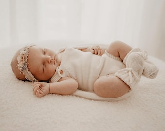 Op de afbeelding: Een slapende baby in een crèmekleurige gebreide romper, bijpassende sokken en een bloemenhoofdband. De baby ligt op een zacht, wit, getextureerd oppervlak. De romper heeft houten knopen.