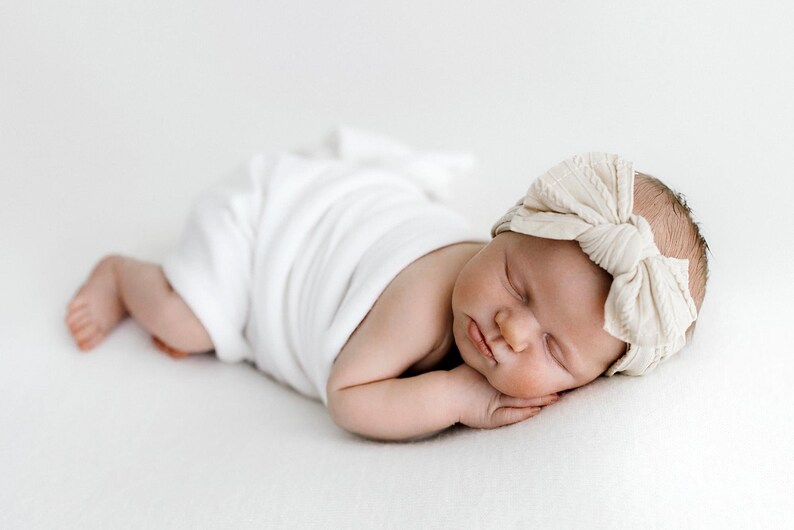 May include: A sleeping infant wrapped in a plain white swaddle blanket, wearing a textured cream-colored headband featuring a large, knotted bow detail on the front. The infant is positioned lying on their stomach with their head resting on hands.
