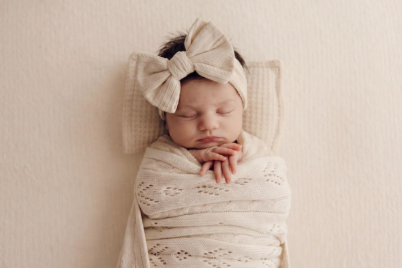 May include: A newborn infant wrapped in a cream-colored knit swaddle blanket with a delicate diamond pattern. The infant wears a matching cream-colored ribbed headband featuring a large, prominent bow centered on the forehead while resting.