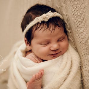 May include: A newborn infant with dark hair is wrapped in a soft, cream-coloured textured blanket. The infant wears a delicate, thin cream-coloured headband adorned with small floral accents. The infant is sleeping peacefully with a slight smile.
